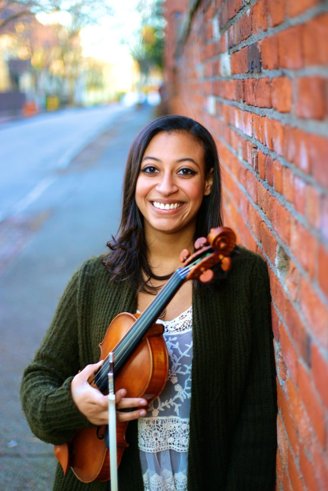 Carmen Johnson Pájaro holding a violin and leaning on a brick wall